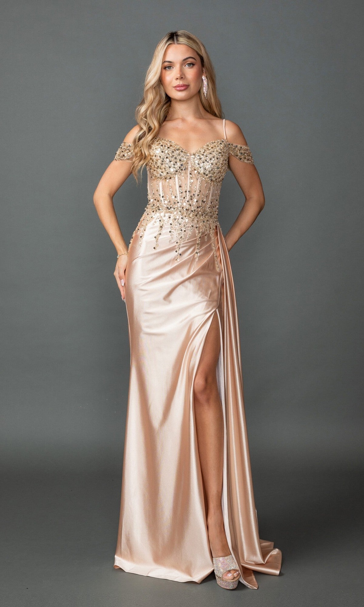 A woman poses against a gray background in the Dancing Queen 4551 long prom dress in champagne, featuring a beaded and sequin embellished bodice, high slit, sheer corset details, and peep-toe heels.